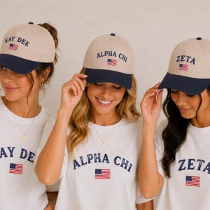May include: Three women wearing matching baseball caps and t-shirts. The caps are tan with navy blue brims and feature text and an American flag. The t-shirts are white with matching text and flags.