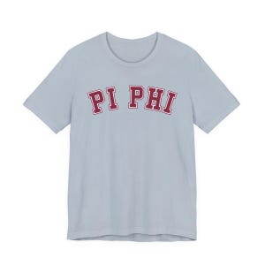 Könnte beinhalten: Hellblaues T-Shirt mit dem Text "PI PHI" in weinroten Blockbuchstaben. Die Buchstaben haben eine weiße Umrandung. Das Shirt hat kurze Ärmel und einen Rundhalsausschnitt.
