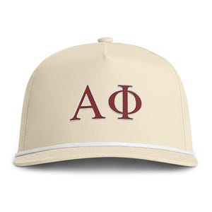 May include: A cream-coloured baseball cap featuring the Greek letters Alpha and Phi embroidered in a dark red thread on the front panel. The cap includes a white cord trim running horizontally across the base of the crown above the brim.