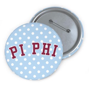 May include: A round pin-back button featuring a light blue background with a white polka dot pattern. The text PI PHI is printed in a maroon collegiate-style font across the center. The back of the button shows a silver metal pin mechanism.