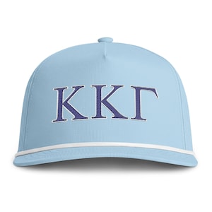 Puede incluir: Una gorra de béisbol azul claro con las letras griegas KKG bordadas en azul oscuro con un borde blanco. La gorra incluye un detalle de cuerda trenzada blanca en la base de la corona, justo encima de la visera curva, y un botón superior.