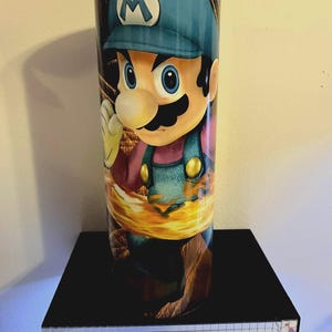 Puede incluir: Un vaso cil&iacute;ndrico con una vibrante ilustraci&oacute;n del personaje de videojuego Mario. El dise&ntilde;o muestra a Mario con su gorra azul y su peto, sobre un fondo de fuego. El vaso se exhibe en una estanter&iacute;a negra.