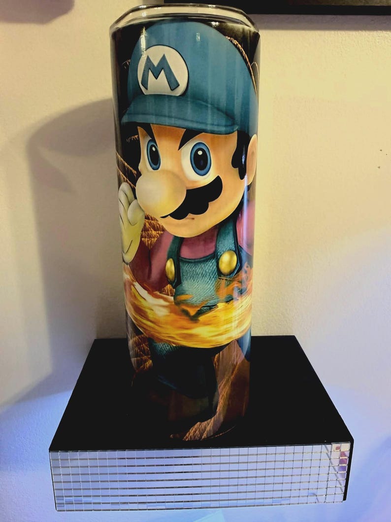 Puede incluir: Un vaso cil&iacute;ndrico con una vibrante ilustraci&oacute;n del personaje de videojuego Mario. El dise&ntilde;o muestra a Mario con su gorra azul y su peto, sobre un fondo de fuego. El vaso se exhibe en una estanter&iacute;a negra.