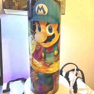 Puede incluir: Un vaso cil&iacute;ndrico alto con una vibrante ilustraci&oacute;n del personaje de videojuego, Luigi. El dise&ntilde;o muestra a Luigi con su mono verde y gorra azul. El vaso est&aacute; sobre un fondo colorido y parece ser de acero inoxidable.