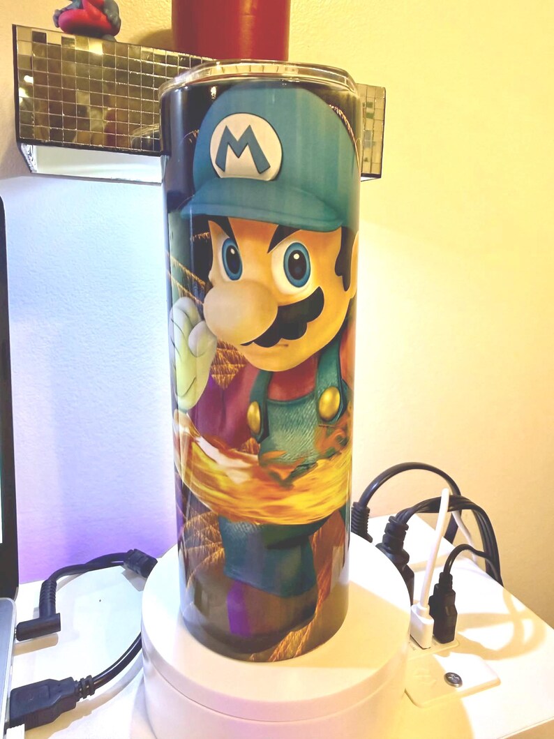 Puede incluir: Un vaso cil&iacute;ndrico alto con una vibrante ilustraci&oacute;n del personaje de videojuego, Luigi. El dise&ntilde;o muestra a Luigi con su mono verde y gorra azul. El vaso est&aacute; sobre un fondo colorido y parece ser de acero inoxidable.
