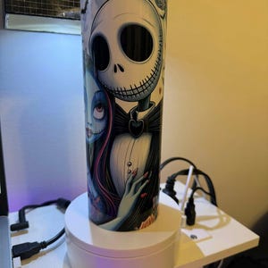 Puede incluir: Un vaso cil&iacute;ndrico con Jack Skellington y Sally de Pesadilla antes de Navidad. El dise&ntilde;o incluye una calavera blanca con pajarita negra y una mujer con pelo largo rojo. El vaso est&aacute; sobre un soporte giratorio blanco.