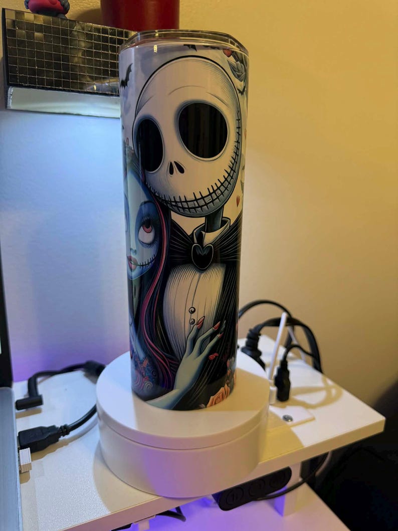 Puede incluir: Un vaso cil&iacute;ndrico con Jack Skellington y Sally de Pesadilla antes de Navidad. El dise&ntilde;o incluye una calavera blanca con pajarita negra y una mujer con pelo largo rojo. El vaso est&aacute; sobre un soporte giratorio blanco.