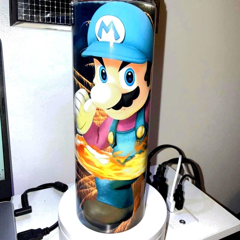 Puede incluir: Un vaso cil&iacute;ndrico alto con una ilustraci&oacute;n de un personaje de dibujos animados con gorra azul y peto. El personaje tiene un gran bigote y est&aacute; rodeado de llamas. El vaso es probablemente para bebidas fr&iacute;as.