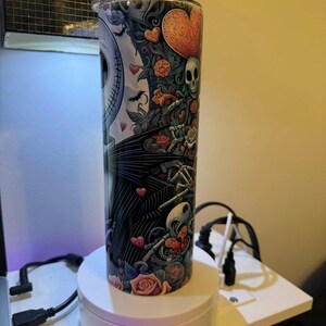 Puede incluir: Un vaso alto de acero inoxidable con un dise&ntilde;o colorido que presenta a Jack Skellington, corazones, rosas y calaveras. El dise&ntilde;o est&aacute; sobre un fondo blanco y negro con una tapa plateada. El vaso es cil&iacute;ndrico y est&aacute; sobre un soporte giratorio blanco.