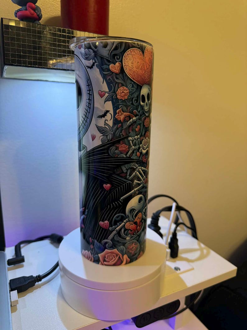 Puede incluir: Un vaso alto de acero inoxidable con un dise&ntilde;o colorido que presenta a Jack Skellington, corazones, rosas y calaveras. El dise&ntilde;o est&aacute; sobre un fondo blanco y negro con una tapa plateada. El vaso es cil&iacute;ndrico y est&aacute; sobre un soporte giratorio blanco.