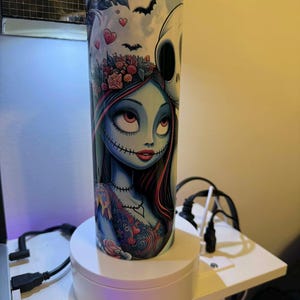 Puede incluir: Un vaso de acero inoxidable con una colorida ilustraci&oacute;n de Sally de Pesadilla antes de Navidad. El dise&ntilde;o incluye murci&eacute;lagos, corazones y Jack Skellington. El vaso es cil&iacute;ndrico y tiene una tapa plateada.