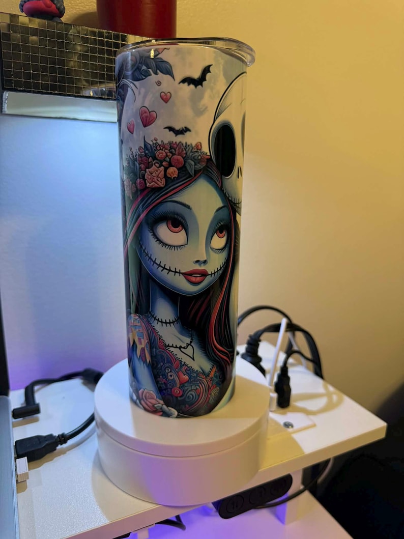 Puede incluir: Un vaso de acero inoxidable con una colorida ilustraci&oacute;n de Sally de Pesadilla antes de Navidad. El dise&ntilde;o incluye murci&eacute;lagos, corazones y Jack Skellington. El vaso es cil&iacute;ndrico y tiene una tapa plateada.