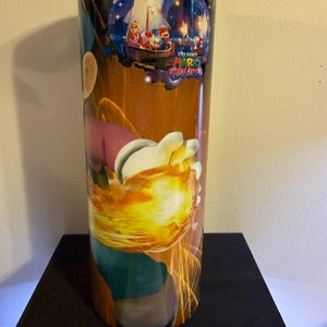 Puede incluir: Un vaso alto y cil&iacute;ndrico con un dise&ntilde;o colorido de "The Super Mario Galaxy". La obra de arte representa a Mario, la Princesa Peach y otros personajes en un barco bajo un cielo estrellado, con un efecto de explosi&oacute;n de fuego.