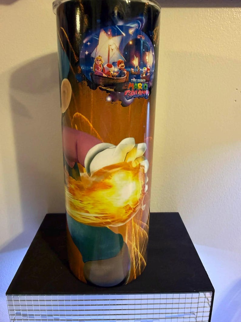 Puede incluir: Un vaso alto y cil&iacute;ndrico con un dise&ntilde;o colorido de "The Super Mario Galaxy". La obra de arte representa a Mario, la Princesa Peach y otros personajes en un barco bajo un cielo estrellado, con un efecto de explosi&oacute;n de fuego.