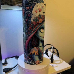 Puede incluir: Un vaso alto y cil&iacute;ndrico con una colorida ilustraci&oacute;n de un personaje con rasgos cosidos, detalles florales y detalles en forma de coraz&oacute;n. El dise&ntilde;o utiliza colores oscuros y claros, y tiene una tapa transparente. El vaso est&aacute; sobre un soporte giratorio blanco.