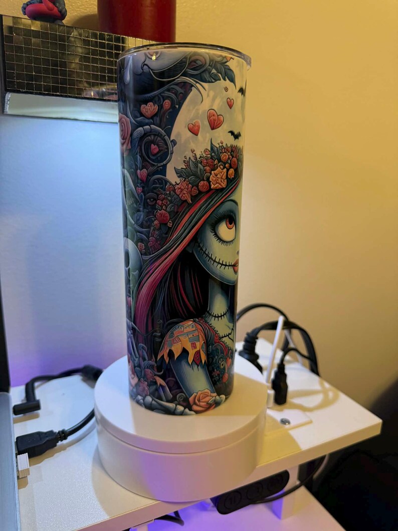 Puede incluir: Un vaso alto y cil&iacute;ndrico con una colorida ilustraci&oacute;n de un personaje con rasgos cosidos, detalles florales y detalles en forma de coraz&oacute;n. El dise&ntilde;o utiliza colores oscuros y claros, y tiene una tapa transparente. El vaso est&aacute; sobre un soporte giratorio blanco.