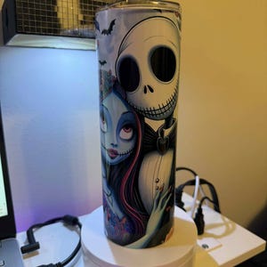 Puede incluir: Vaso cil&iacute;ndrico de acero inoxidable con una ilustraci&oacute;n de Jack Skellington y Sally de Pesadilla antes de Navidad. El dise&ntilde;o incluye murci&eacute;lagos y una est&eacute;tica oscura y caprichosa. El vaso tiene una tapa plateada.