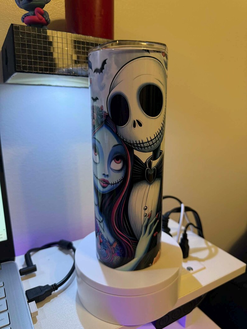 Puede incluir: Vaso cil&iacute;ndrico de acero inoxidable con una ilustraci&oacute;n de Jack Skellington y Sally de Pesadilla antes de Navidad. El dise&ntilde;o incluye murci&eacute;lagos y una est&eacute;tica oscura y caprichosa. El vaso tiene una tapa plateada.