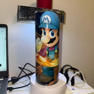 Puede incluir: Un vaso cil&iacute;ndrico con una vibrante ilustraci&oacute;n de Mario del videojuego Super Mario Bros. El dise&ntilde;o muestra a Mario con su gorra azul y su mono, sobre un fondo de fuego. El vaso est&aacute; colocado sobre un soporte giratorio blanco.