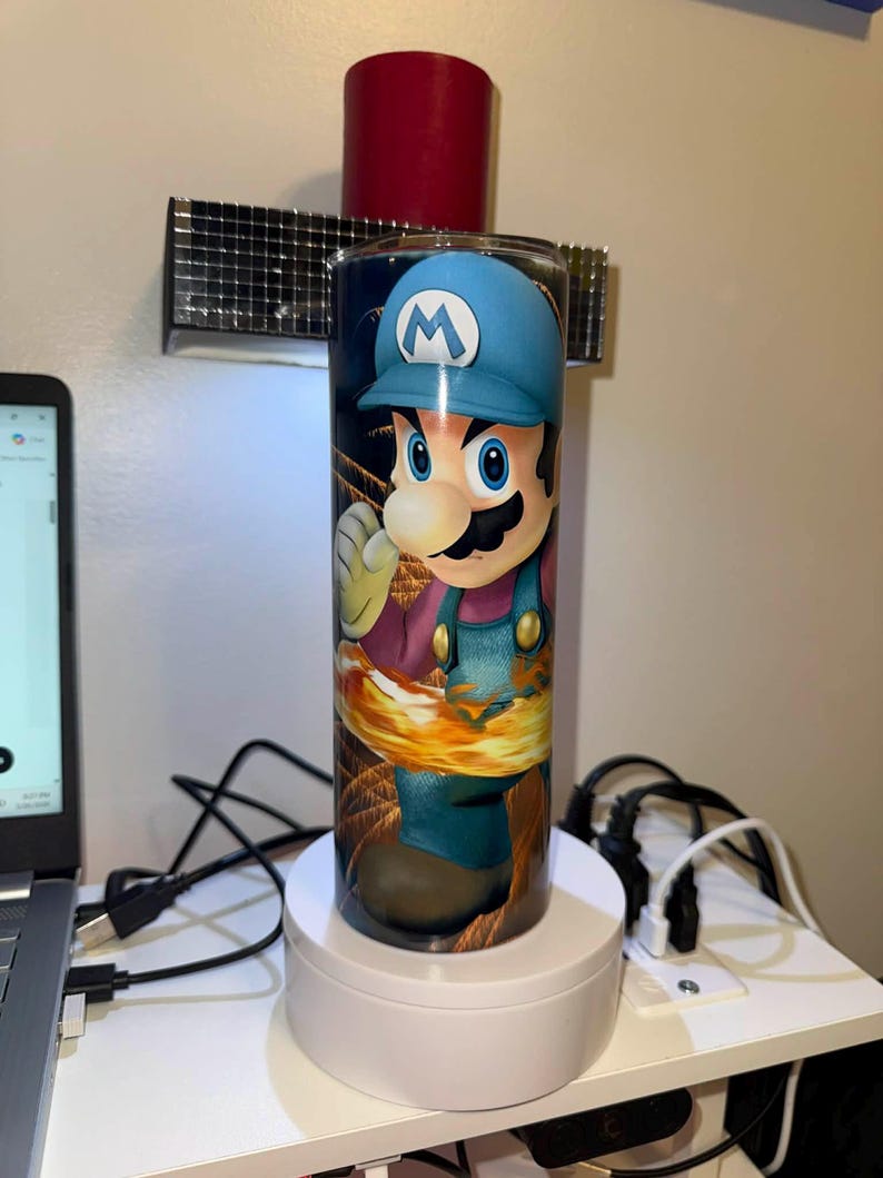 Puede incluir: Un vaso cil&iacute;ndrico con una vibrante ilustraci&oacute;n de Mario del videojuego Super Mario Bros. El dise&ntilde;o muestra a Mario con su gorra azul y su mono, sobre un fondo de fuego. El vaso est&aacute; colocado sobre un soporte giratorio blanco.