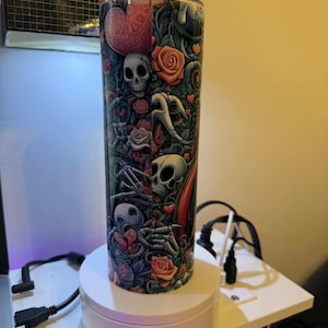 Puede incluir: Un vaso alto y cil&iacute;ndrico con un dise&ntilde;o colorido que presenta calaveras, rosas, corazones y figuras esquel&eacute;ticas. La obra de arte es predominantemente en tonos de rojo, gris y negro, creando un tema g&oacute;tico o rom&aacute;ntico. El vaso se exhibe en un soporte giratorio blanco.