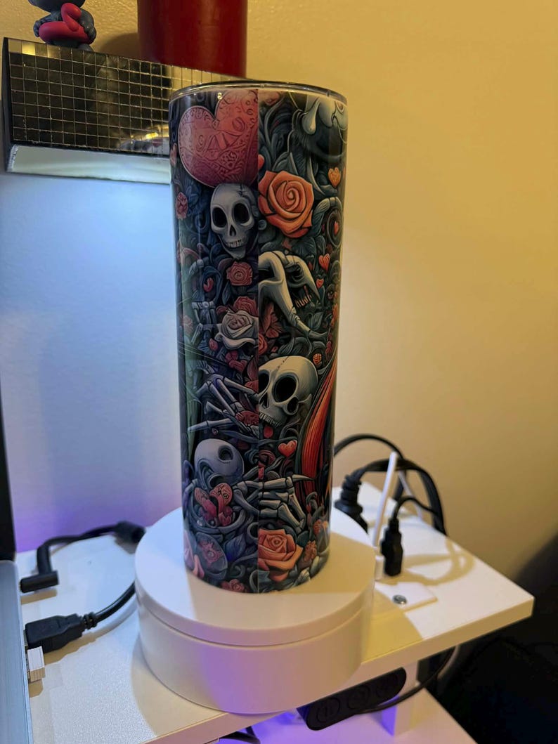 Puede incluir: Un vaso alto y cil&iacute;ndrico con un dise&ntilde;o colorido que presenta calaveras, rosas, corazones y figuras esquel&eacute;ticas. La obra de arte es predominantemente en tonos de rojo, gris y negro, creando un tema g&oacute;tico o rom&aacute;ntico. El vaso se exhibe en un soporte giratorio blanco.