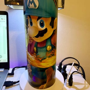 Puede incluir: Un vaso cil&iacute;ndrico con un gr&aacute;fico vibrante de un personaje de dibujos animados con un gorro azul y un peto. El personaje tiene bigote y est&aacute; sobre un fondo de llamas. El vaso es probablemente de acero inoxidable y est&aacute; dise&ntilde;ado para bebidas fr&iacute;as o calientes.