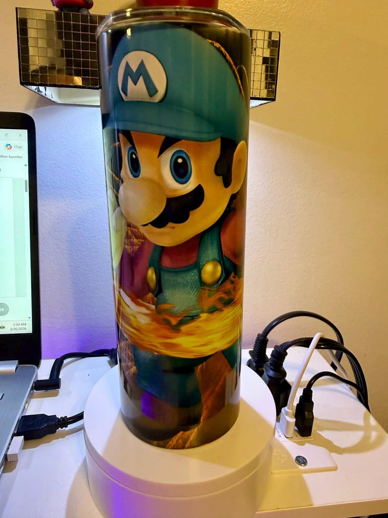 Puede incluir: Un vaso cil&iacute;ndrico con un gr&aacute;fico vibrante de un personaje de dibujos animados con un gorro azul y un peto. El personaje tiene bigote y est&aacute; sobre un fondo de llamas. El vaso es probablemente de acero inoxidable y est&aacute; dise&ntilde;ado para bebidas fr&iacute;as o calientes.