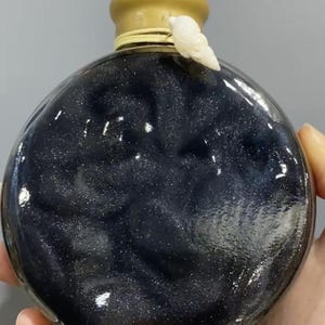 Puede incluir: Una botella redonda de vidrio transparente llena de un líquido azul oscuro y brillante. La botella tiene una tapa dorada y un pequeño adorno de concha blanca. La botella se sostiene en una mano.