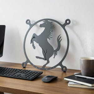 May include: A gray metal wall art piece featuring a prancing horse within a circular frame. The art piece is displayed on a wooden desk, alongside a keyboard, mouse, and a coffee mug. The word "Ferrari" is visible on the art piece.