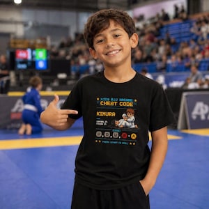 May include: Black t-shirt with a graphic that reads "KIDS BJJ ARCADE: CHEAT CODE" and "KIMURA". The graphic includes a cartoon illustration of a martial artist and a series of button codes. The shirt is worn by a smiling child.
