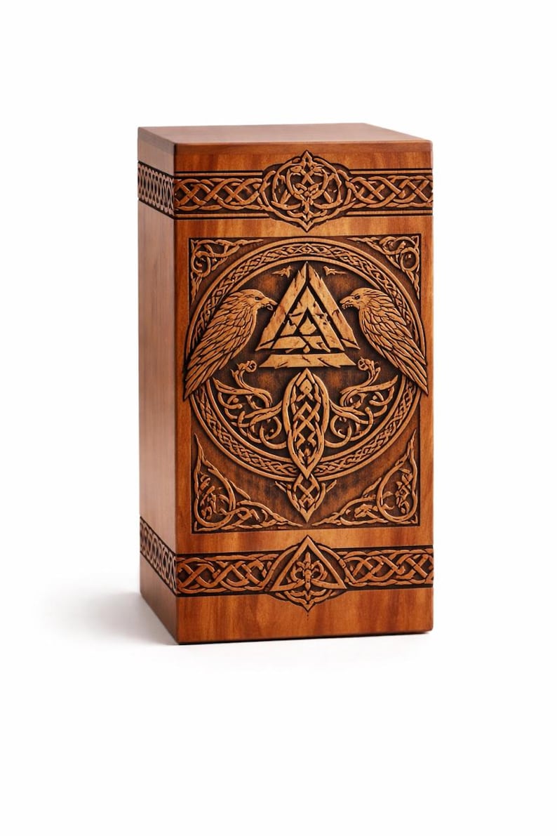 K&ouml;nnte beinhalten: Eine rechteckige Holzbox mit aufwendigen Schnitzereien auf der Vorderseite. Das Design zeigt zwei Raben, die ein zentrales Valknut-Symbol in einem kreisf&ouml;rmigen Rahmen flankieren, umgeben von detaillierten keltischen Knotenmustern an den R&auml;ndern.