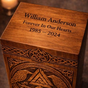 K&ouml;nnte beinhalten: Eine rechteckige Urne aus Holz mit dunkelbrauner Oberfl&auml;che und detaillierten Schnitzereien von zwei Raben, die ein zentrales dreieckiges Knotenmuster flankieren. Auf der Oberseite ist der Text William Anderson, Forever In Our Hearts, 1985-2024 eingraviert.