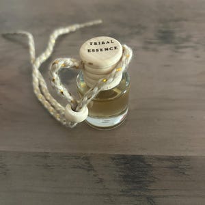Könnte beinhalten: Kleine Glasflasche gefüllt mit einer goldenen Flüssigkeit, bedeckt mit einem Holzdeckel mit der Aufschrift "TRIBAL ESSENCE". Ein cremefarbener Faden mit goldenen Akzenten ist befestigt. Auf einer Holzoberfläche platziert.