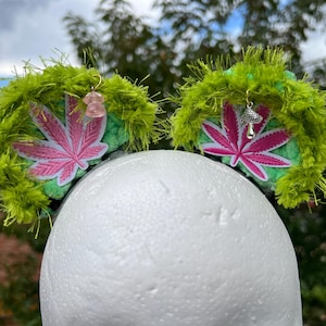 May include: A headband featuring two round ears covered in bright green fuzzy yarn. Each ear displays a pink cannabis leaf patch. A small pink translucent mushroom charm hangs from the left ear, and a silver mushroom charm hangs from the right.