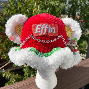 May include: A bright red crocheted bucket hat featuring a white faux fur brim with green accents. The hat displays a rectangular patch with the text Effin in silver, a silver chain draped across the front, and two large white fuzzy ears.