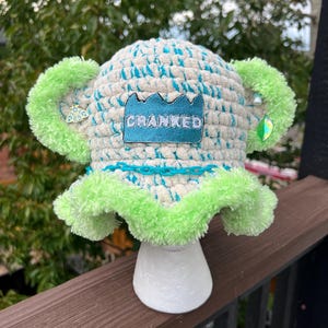 May include: A crochet bucket hat featuring a cream and teal variegated yarn body with a bright lime green fuzzy brim and two matching fuzzy ears. A blue patch with the text CRANKED is centered on the front, accented by a teal plastic chain.