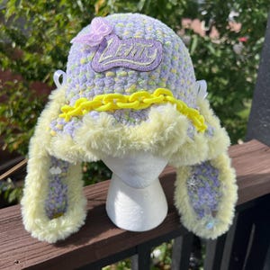May include: A lavender and yellow variegated crochet hat with long ear flaps, trimmed in yellow faux fur. The hat features a bright yellow plastic chain across the front, a purple embroidered patch reading LEVITY, and a small light pink butterfly accent.