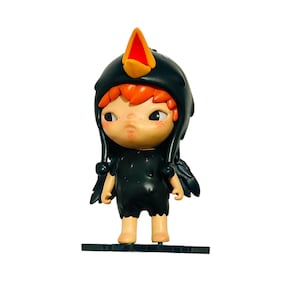 May include: A figurine of a child dressed in a black bird costume with a yellow and red beak. The figure has red hair, a pale complexion, and is standing on a black base. The costume includes small wing details.