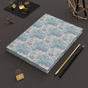 May include: A rectangular notebook featuring a repeating pattern of light blue pumpkins, small flowers, and intricate gray foliage on a white background. The notebook is positioned alongside two pencils, one gold and one black, and several gold binder clips.