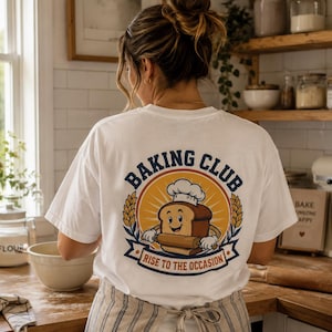 Könnte beinhalten: Ein weißes T-Shirt mit einer Grafik auf der Rückseite, die eine Cartoon-Brotscheibe mit Kochmütze zeigt, die ein Nudelholz hält. Das Design enthält den Text BAKING CLUB und RISE TO THE OCCASION in Blau und Orange mit Weizenähren.