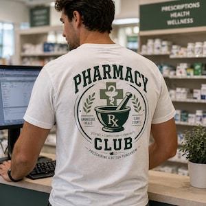 Könnte beinhalten: Weißes T-Shirt mit einem grün-schwarzen Pharmacy Club-Design auf dem Rücken. Das Design umfasst einen Mörser und Stößel, ein medizinisches Kreuz und die Worte "Knowledge Heals" und "Care Counts". Das Shirt hat auch die Worte "Prescribing a Better Tomorrow".