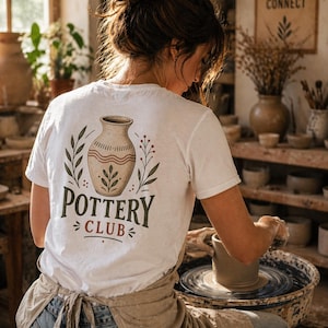 May include: A person wearing a white t-shirt with a pottery design and the words "POTTERY CLUB". The person is working on a pottery wheel, creating a clay pot. The scene is set in a pottery studio with natural light.