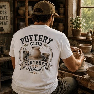 Könnte beinhalten: Ein weißes T-Shirt mit einem Aufdruck auf der Rückseite, der den Text Pottery Club Centered on the Wheel zeigt. Das Design umfasst eine Illustration eines Tontopfes auf einer Töpferscheibe, einen Becher mit Werkzeugen und eine kleine Schale.