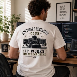 Könnte beinhalten: Ein weißes T-Shirt mit einem Rückendruck, der eine Person an einem Schreibtisch mit drei Computermonitoren zeigt. Der Text lautet "SOFTWARE DEVELOPER CLUB" und "IT WORKS ON MY MACHINE", ergänzt durch Codesymbole und eine kleine Topfpflanze.