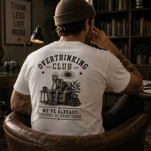 Könnte beinhalten: Ein weißes T-Shirt mit einer Grafik auf der Rückseite, die ein Skelett in einem Sessel mit einer Gedankenblase, einem Beistelltisch und einer Pflanze zeigt. Der Text lautet OVERTHINKING CLUB und WE'VE ALREADY THOUGHT OF EVERYTHING in Schwarz.