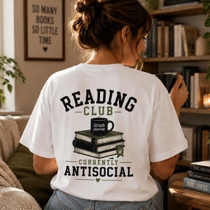Könnte beinhalten: Ein weißes Kurzarm-T-Shirt mit einem Rückendruck, der den Text READING CLUB und CURRENTLY ANTISOCIAL in Schwarz und Olivgrün zeigt. Das Motiv besteht aus einem Bücherstapel, auf dem eine schwarze Tasse mit der Aufschrift ONE MORE CHAPTER steht.