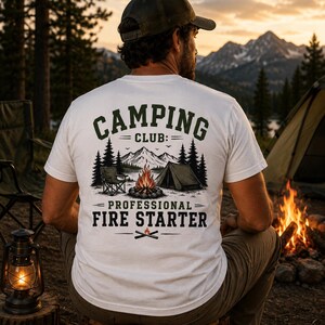 Könnte beinhalten: Ein weißes T-Shirt mit einem Aufdruck auf der Rückseite, der den Text CAMPING CLUB PROFESSIONAL FIRE STARTER zeigt. Das Motiv umfasst ein Lagerfeuer, ein Zelt, einen Klappstuhl und eine Berglandschaft mit Tannen in dunkelgrüner und schwarzer Farbe.