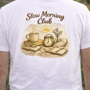 Op de afbeelding: Wit T-shirt met een afbeelding van een koffiekopje, boeken, een wekker en de tekst "Slow Morning Club". Het ontwerp bevat een zonsopgang en een kleine vaas met groen. Het shirt is een casual kledingstuk.