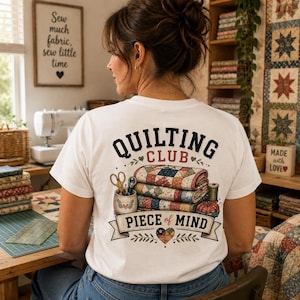 Può includere: Una t-shirt bianca con una grafica sul retro che mostra una pila di trapunte colorate a motivi, forbici in un contenitore, un rocchetto di filo e un puntaspilli. Il testo recita QUILTING CLUB PIECE of MIND con decorazioni a cuore e foglie.